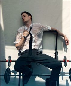 a man in white shirt and tie leaning against wall with barbells on it