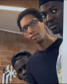 two men standing next to each other in front of a brick wall and wearing glasses