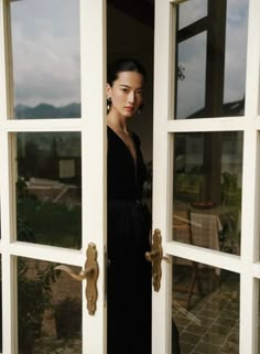 a woman in a black dress standing at the entrance to a house with large windows