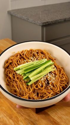 a person holding a bowl of noodles with cucumbers and sesame seeds on top