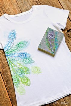 a white t - shirt with peacock feathers painted on it and a book laying next to it