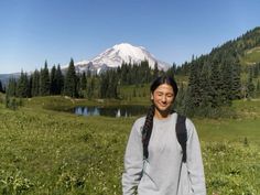 a woman standing in front of a mountain with trees and grass on the ground, smiling at the camera