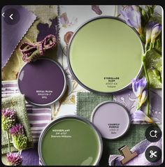 several shades of green and purple paint with flowers on the table next to them,