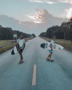 two young men holding surfboards on the side of an empty road in front of trees