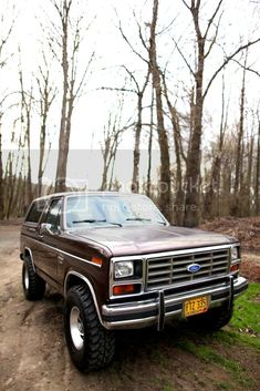 an old ford bronco is parked on the side of a dirt road in front of some trees