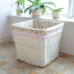 a white basket sitting on the floor next to a window with potted plants in it