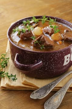 a bowl of beef stew with potatoes and parsley on a napkin next to two spoons