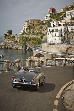 an old car is driving down the road by the water in front of some buildings