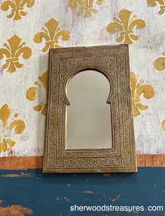 a small mirror sitting on top of a wooden table next to a wallpaper covered with gold florets