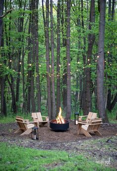 a fire pit in the middle of a forest with chairs around it and string lights hanging from the trees