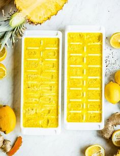 two ice trays filled with yellow liquid next to sliced lemons and pineapples