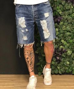 a man with tattoos standing in front of a green wall wearing ripped denim shorts and white sneakers