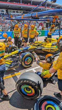 several people in yellow shirts and helmets working on a race car at a track with spectators watching