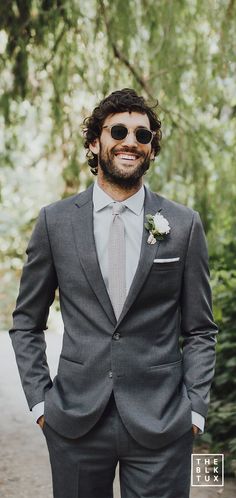 a man in a gray suit and sunglasses smiles at the camera while standing next to some trees