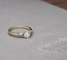 a close up of a diamond ring on a gray surface with an engraved logo in the background