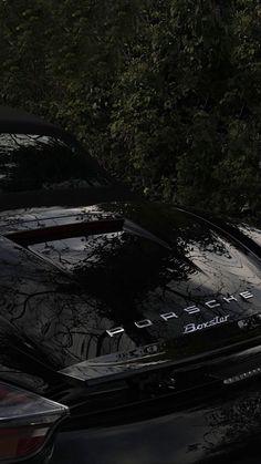 the back end of a black sports car parked in front of some trees and bushes