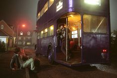 a man standing on top of a purple double - decker bus in the street at night