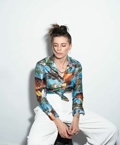 a woman sitting on top of a stool wearing white pants and a shirt with colorful designs