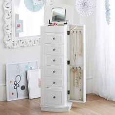 a white jewelry cabinet sitting on top of a hard wood floor next to a mirror