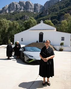 a woman standing in front of a white house next to a sports car with mountains in the background