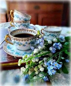 tea cups and saucers on a table with blue flowers in the foreground, next to a book