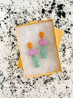 a pair of pink and green earrings sitting on top of a white box with black speckles