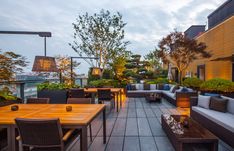 an outdoor seating area with wooden tables and couches, surrounded by greenery at dusk