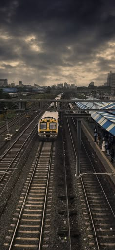 a yellow and white train traveling down tracks under a cloudy sky with buildings in the background