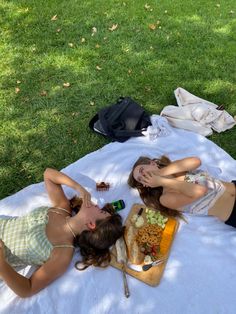 two women laying on a blanket in the grass eating food and drinking beer while another woman sits next to her