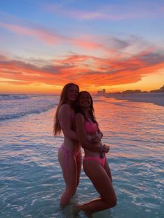 two women in bikinis are standing in the water at sunset, one is hugging the other