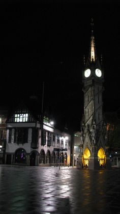 a clock tower lit up at night in the middle of a town square with buildings and cobblestones