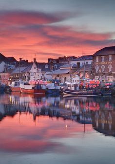 the boats are docked in the harbor at night time, with colorful clouds and buildings reflected in the water