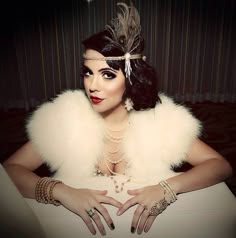 a woman with dark hair and white fur collar sitting at a table in front of a window