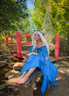 a woman dressed as a fairy sitting on a stone bench next to a stream and bridge