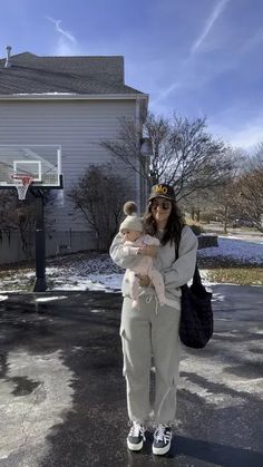 a woman holding a baby standing in front of a basketball hoop on the side of a house