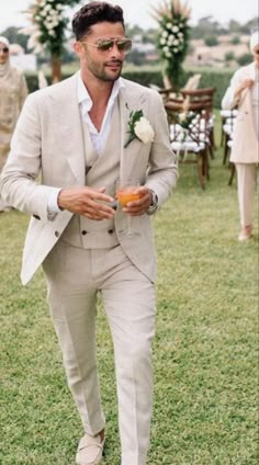 a man in a suit and sunglasses is walking through the grass with an orange drink