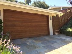 a house with a wooden garage door in front of it and flowers around the driveway