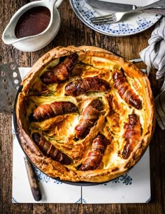 a baked dish with sausages in it on a wooden table next to silverware and utensils