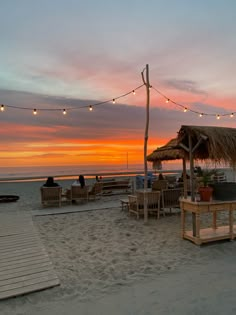 people are sitting at tables on the beach with lights strung over them and thatched umbrellas