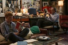 two men sitting on couches reading books in a living room with many other furniture