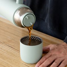 a person pouring coffee into a cup on top of a wooden table next to a metal strainer