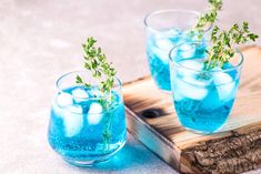 three glasses filled with blue liquid and green sprigs on top of a wooden tray