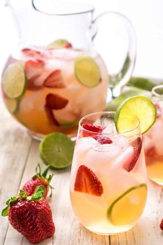 two pitchers of lemonade with strawberries and limes next to them on a wooden table