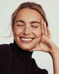 a woman smiling with her eyes closed and hands on her head, wearing a black turtle neck sweater