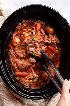 a person holding a fork in a slow cooker filled with meat and vegetables