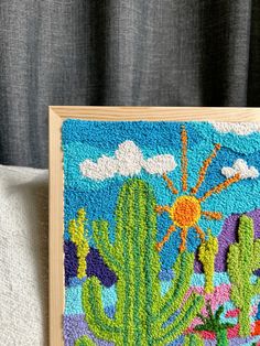 an embroidered cactus scene is displayed on a wooden frame in front of a gray curtain