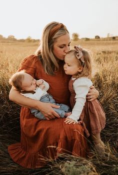 a woman sitting in the grass with two small children and kissing her cheek while she is wearing an orange dress