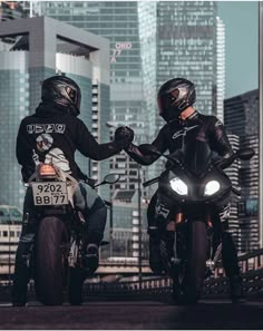 two motorcyclists in leather jackets on their bikes with city buildings in the background