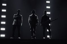 three men are standing on stage with their arms crossed and one is holding a guitar