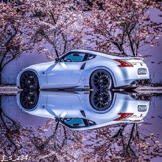 a white sports car is parked in front of some cherry blossom trees with its reflection on the water